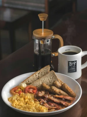 Prato com ovos mexidos, torradas e tomate ao lado de uma prensa de café