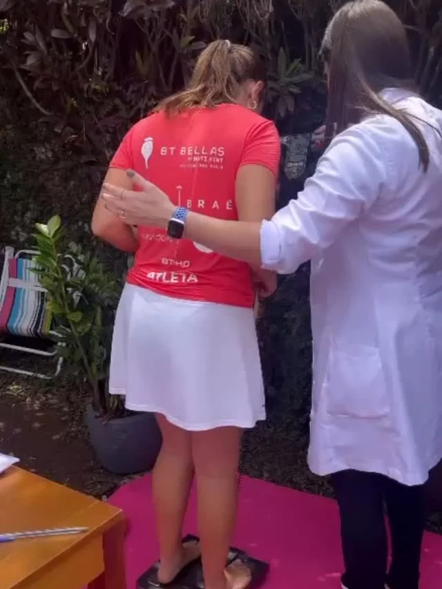 Débora de jaleco ajudando uma mulher a subir na balança de bioimpedância num evento